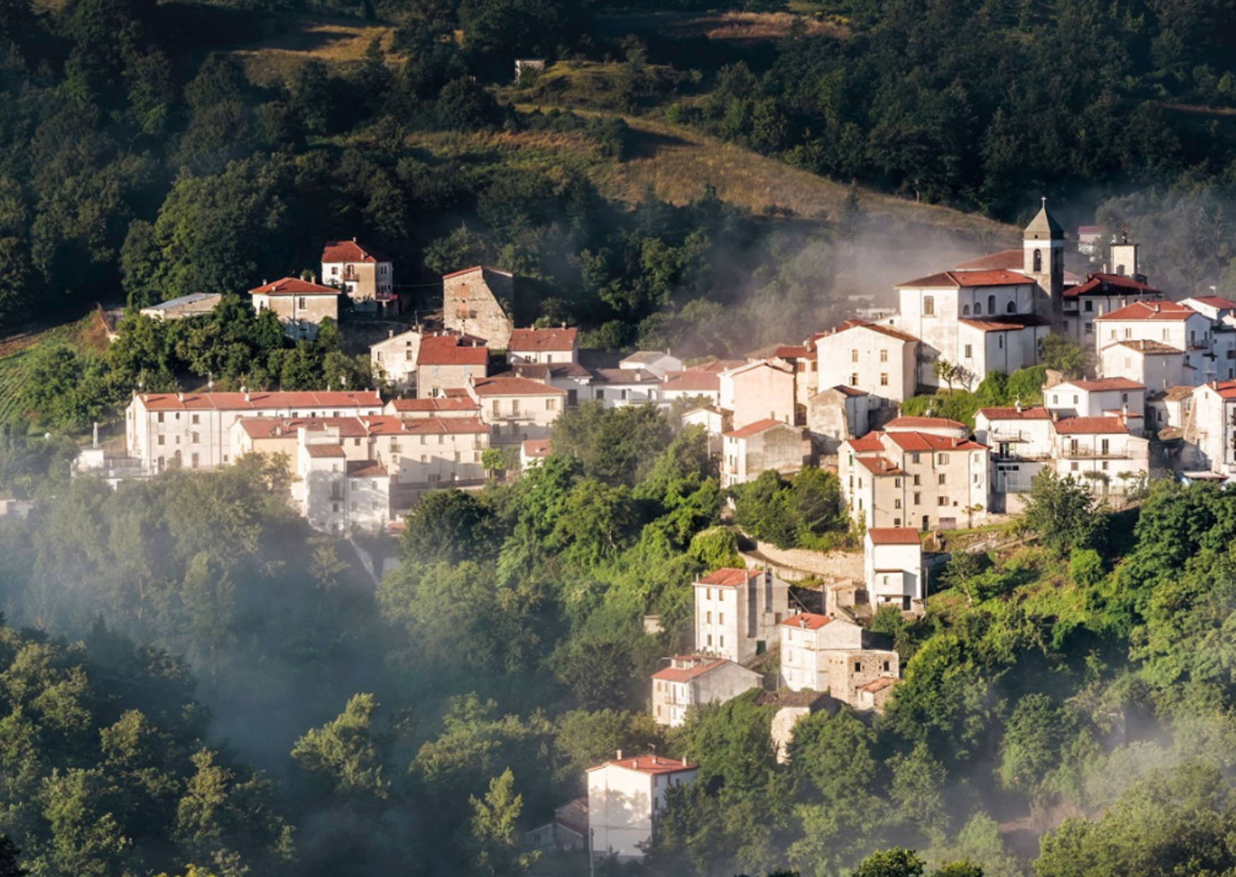 Case chiare e campanile di Castel del Giudice sul versante boscoso, con la nebbia bassa tra gli alberi