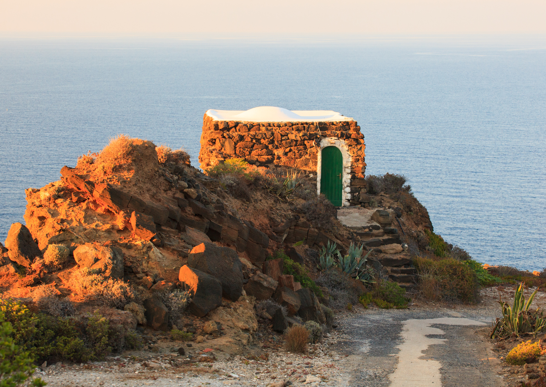 Un dammuso in pietra lavica con porta verde e tetto a volta bianco su una scogliera di Pantelleria