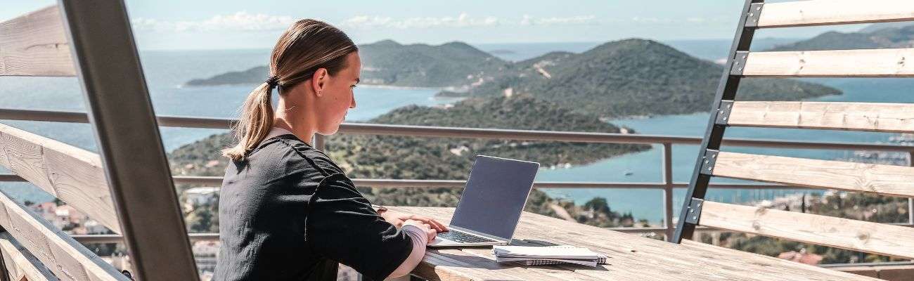 Donna al portatile su una terrazza in legno con vista su una baia: dove vivere in Italia da nomade digitale
