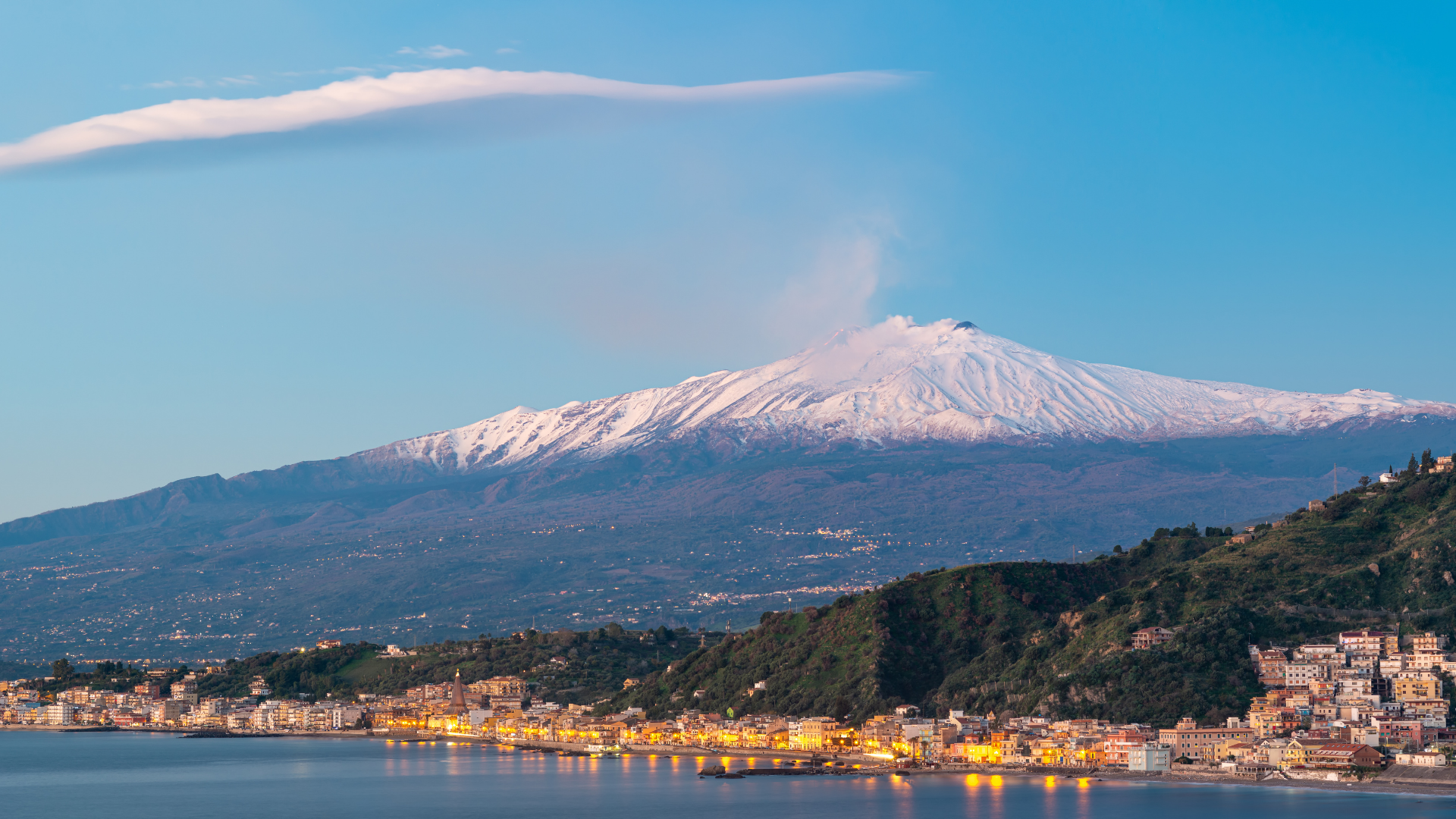 Etna innevato con pennacchio di fumo sopra la costa ionica illuminata al crepuscolo