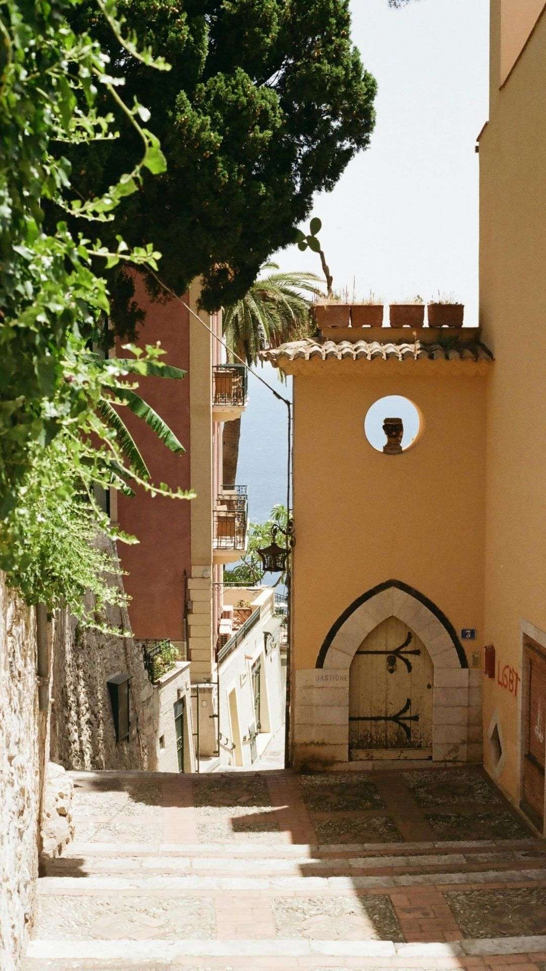 Steinfassade mit Rundfenster und Spitzbogentor in einer begrünten Gasse von Gangi