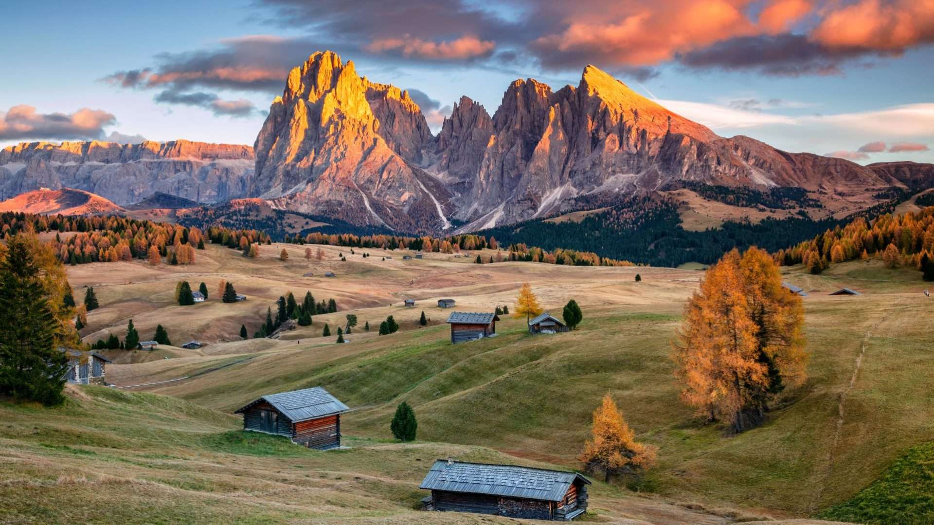 Cumbres de los Dolomitas al atardecer sobre prados alpinos con cabañas de madera y alerces otoñales